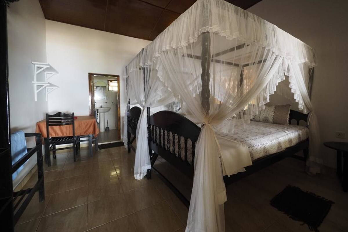 A bedroom with a four-poster canopy bed, a dining table with chairs, and a small table with a lamp.