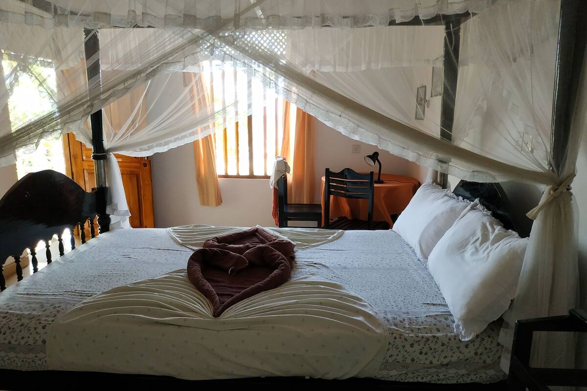 A neatly made bed with a canopy, a chair, and a small table in a room with a window.