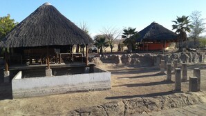 A thatched-roof structure with a concrete foundation, surrounded by a dry, sandy area with sparse vegetation.