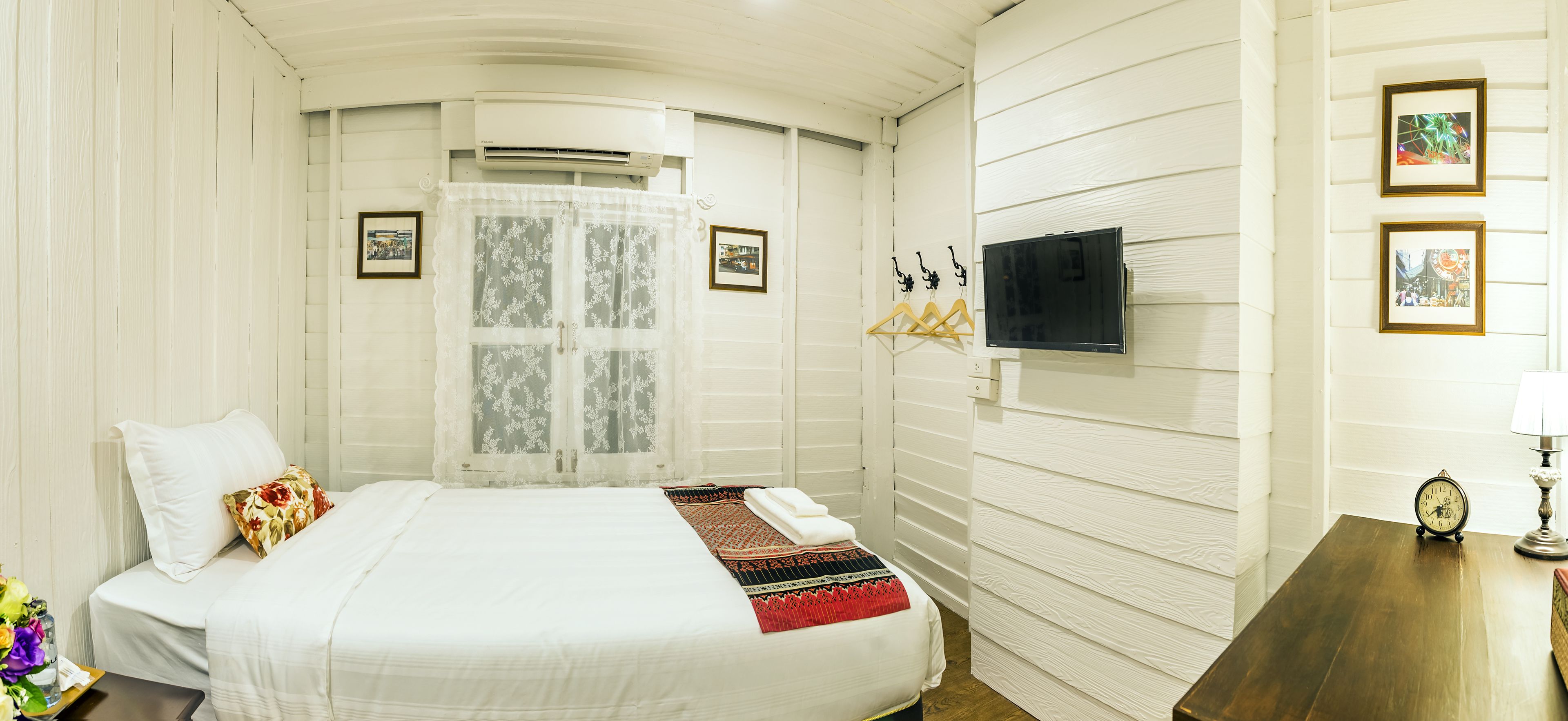 Una camera d'albergo con un letto, una TV montata a parete, una finestra con tenda e un comodino in legno con una lampada.