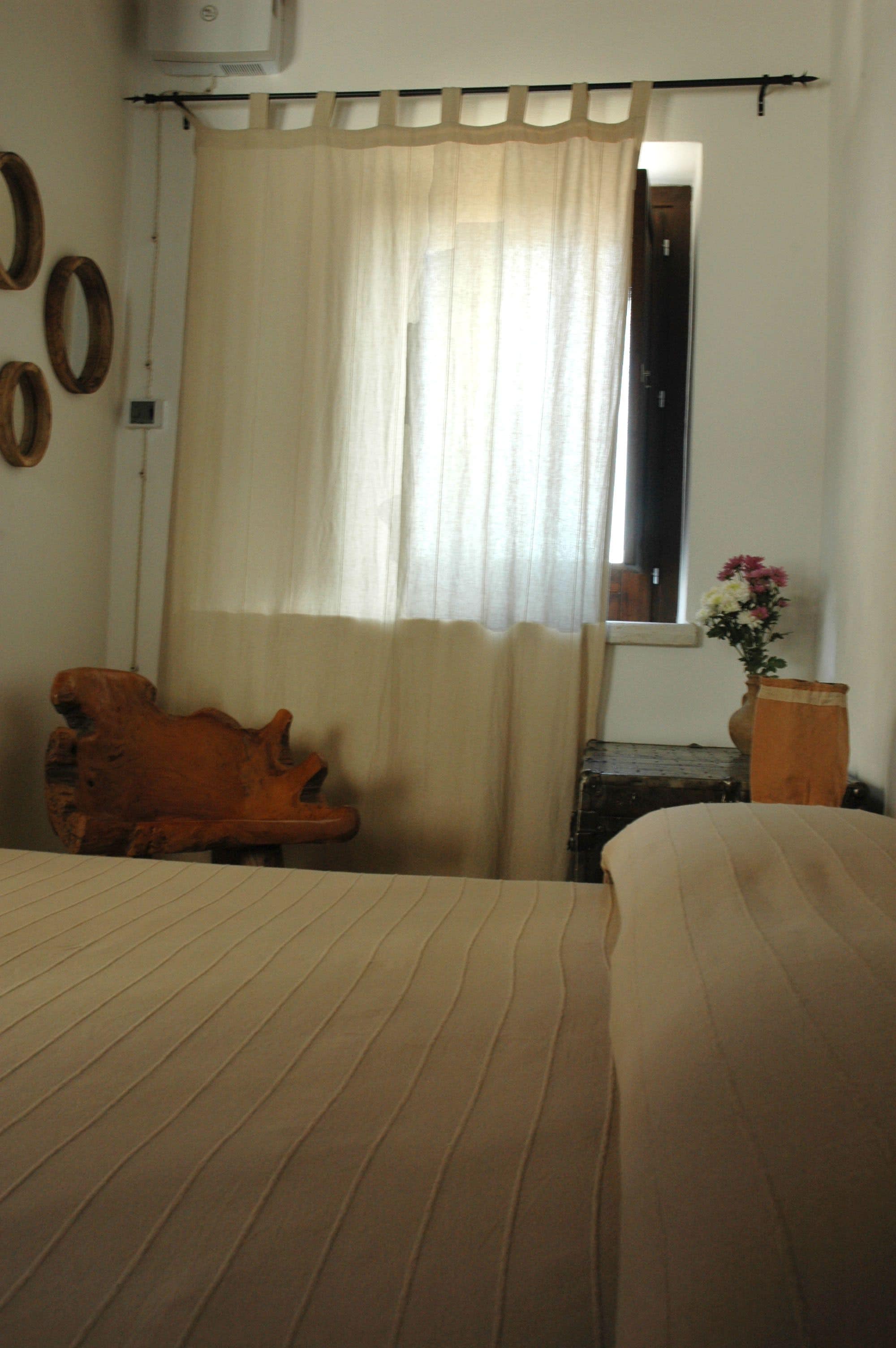 Ein Zimmer mit einem Bett, einem Holzstuhl, einem Fenster mit Vorhang und einer Tischvase mit Blumen.