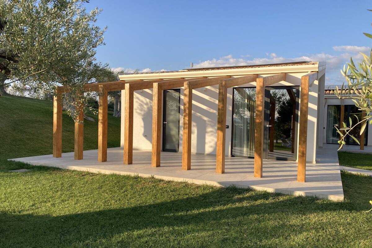 A modern outdoor structure with a wooden pergola and glass doors, surrounded by a well-maintained lawn and trees.