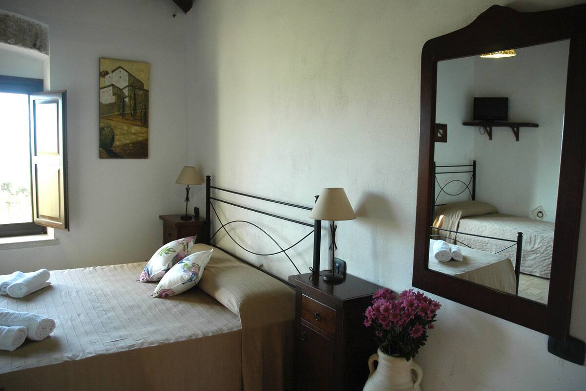 A hotel room with a bed, two bedside lamps, a mirror, a painting on the wall, and a vase with flowers.