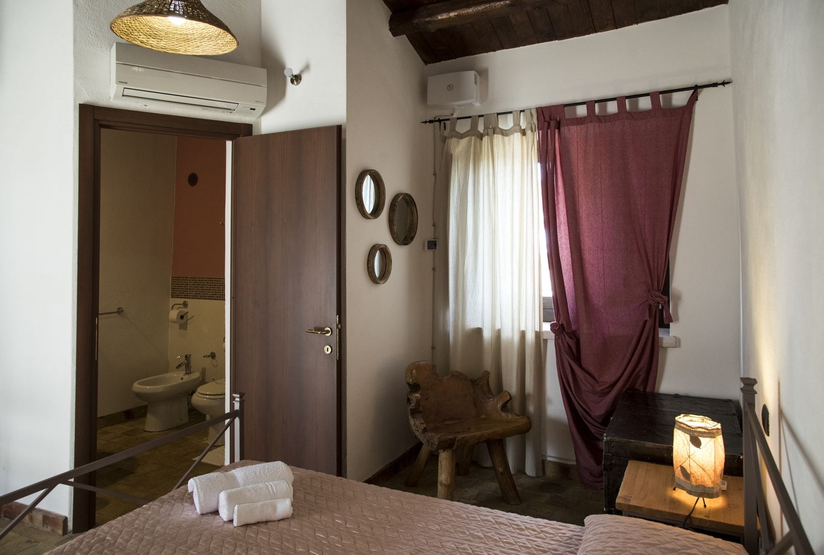 Un dormitorio con cama, una silla de madera, una mesita de noche con lámpara, un baño visible a través de una puerta abierta y una ventana con cortinas.