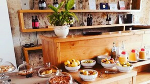 Een buffetopstelling met diverse etenswaren en drankjes, een kamerplant en wijnflessen op planken in de achtergrond.