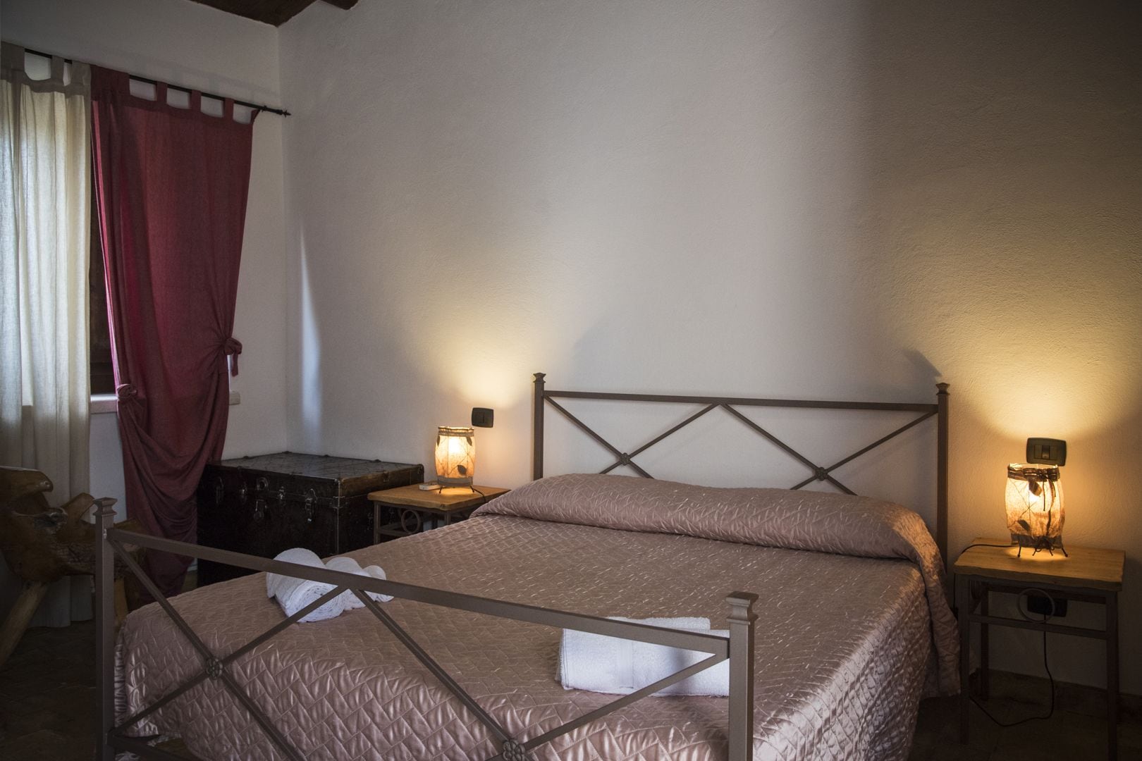 A bedroom with a bed, two bedside tables, a nightstand, a wooden chest, and a window with curtains.