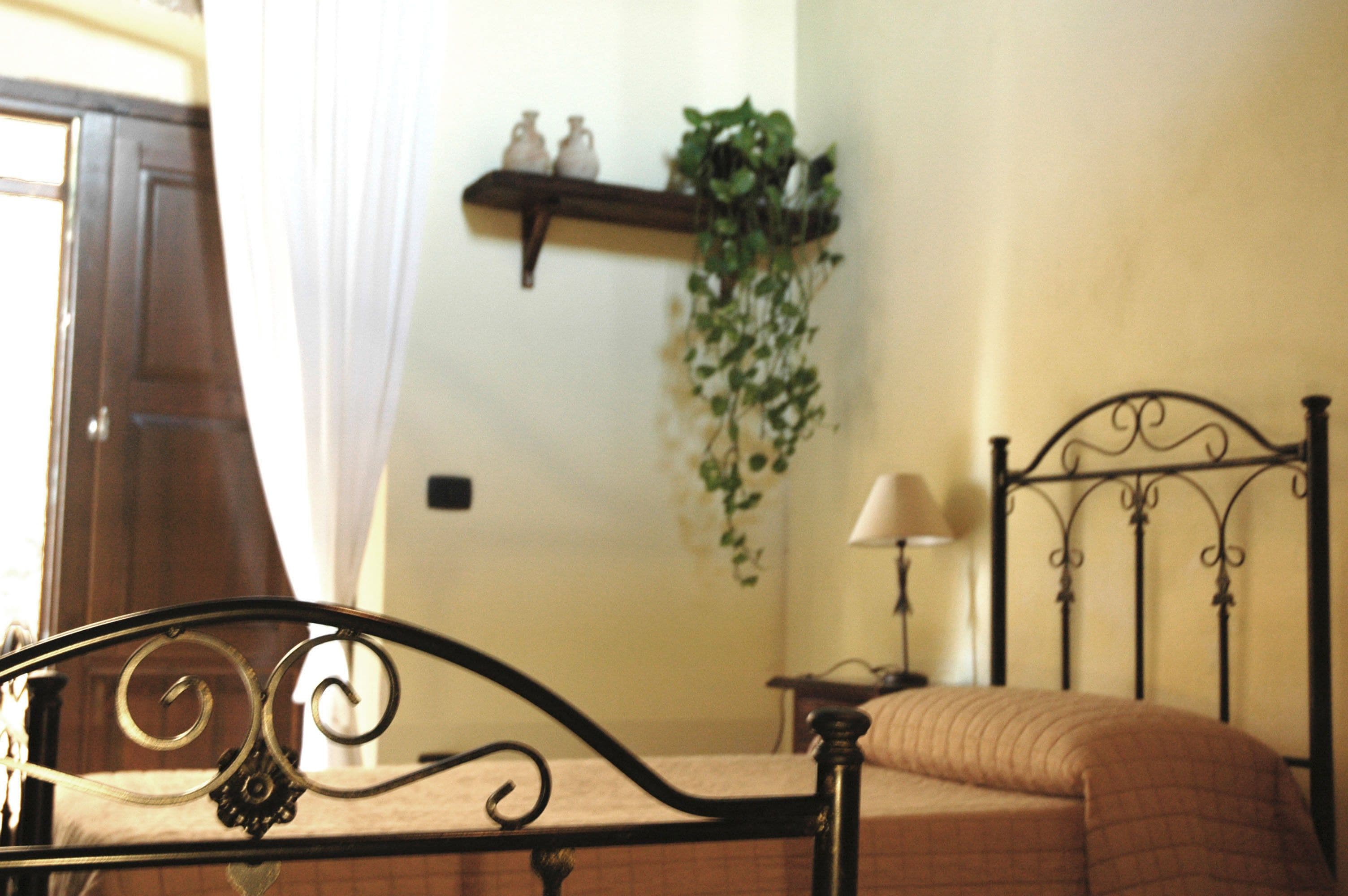 A bedroom with a bed, a bedside table, a lamp, a shelf with decorative items, and a hanging plant.