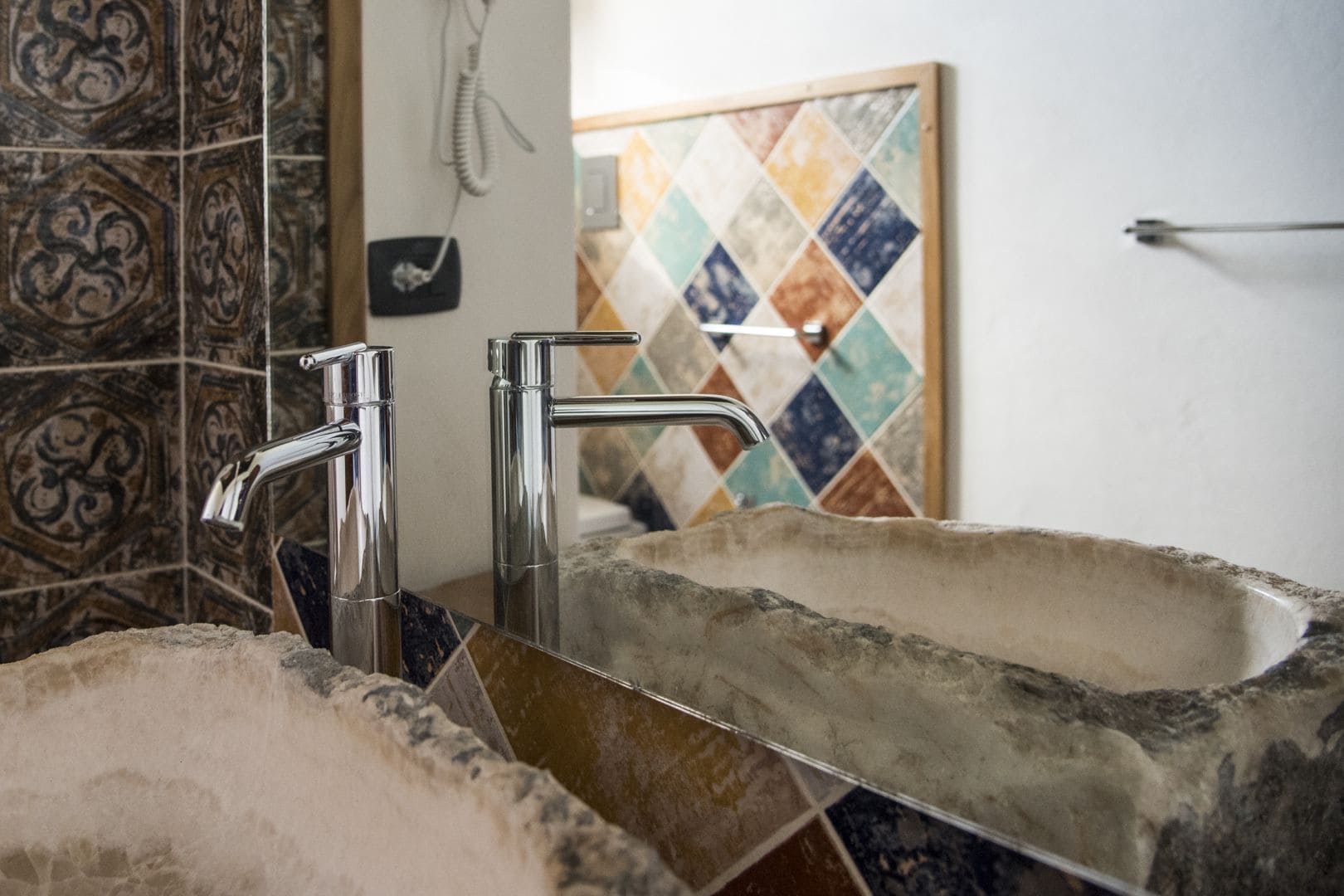 Un baño con un lavamanos de piedra, dos grifos cromados y un salpicadero de mosaico colorido.