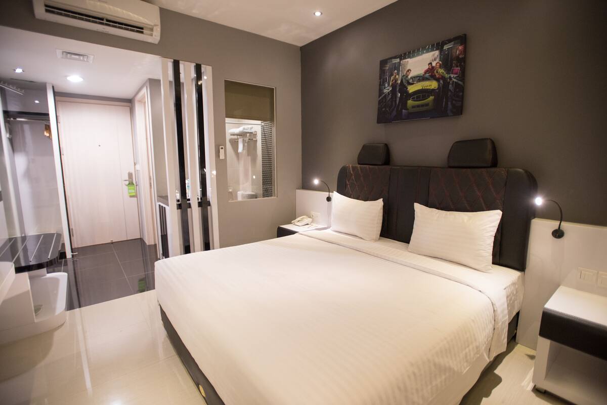 A modern hotel room with a large bed, a bedside table, a nightstand, a mirror, and a painting on the wall.