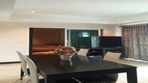 Habitación de hotel moderna con zona de comedor que incluye una mesa negra, sillas blancas, un jarrón de flores, un televisor de pantalla plana y una cama con cortina verde.