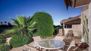 A patio with a round glass table and chairs, surrounded by well-maintained greenery and a view of a building.