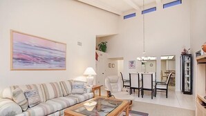 A living room with a sofa, coffee table, and a painting on the wall. There is a dining area with a table and chairs in the background.