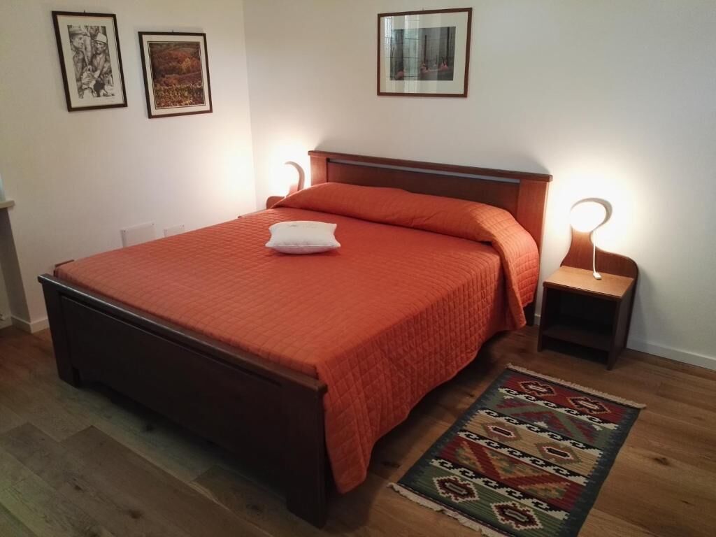 Una cama bien tendida con una colcha roja, dos mesitas de noche con lámparas y una alfombra estampada sobre un piso de madera.