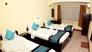A hotel room with three beds, each with a towel and pillow. There is a small table with a bottle and a cup, and a TV mounted on the wall. - Bag Packer's Lodge (Kathmandu)