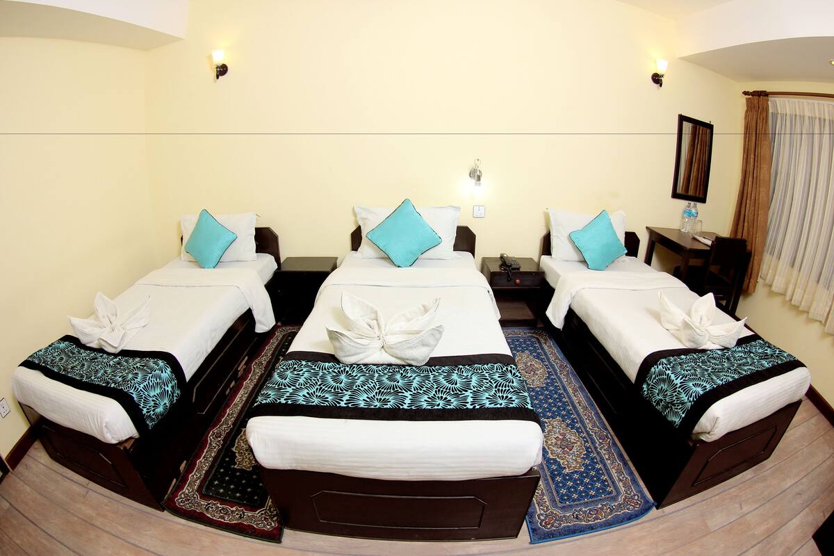 A hotel room with three beds, each with a white bedspread and a blue pillow. There are two bedside tables with lamps, a large mirror, and a window with curtains.