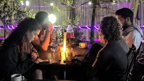 A group of people seated around a table with drinks and a coffee cup, in an indoor setting with plants and string lights. - Bag Packer's Lodge (Kathmandu)