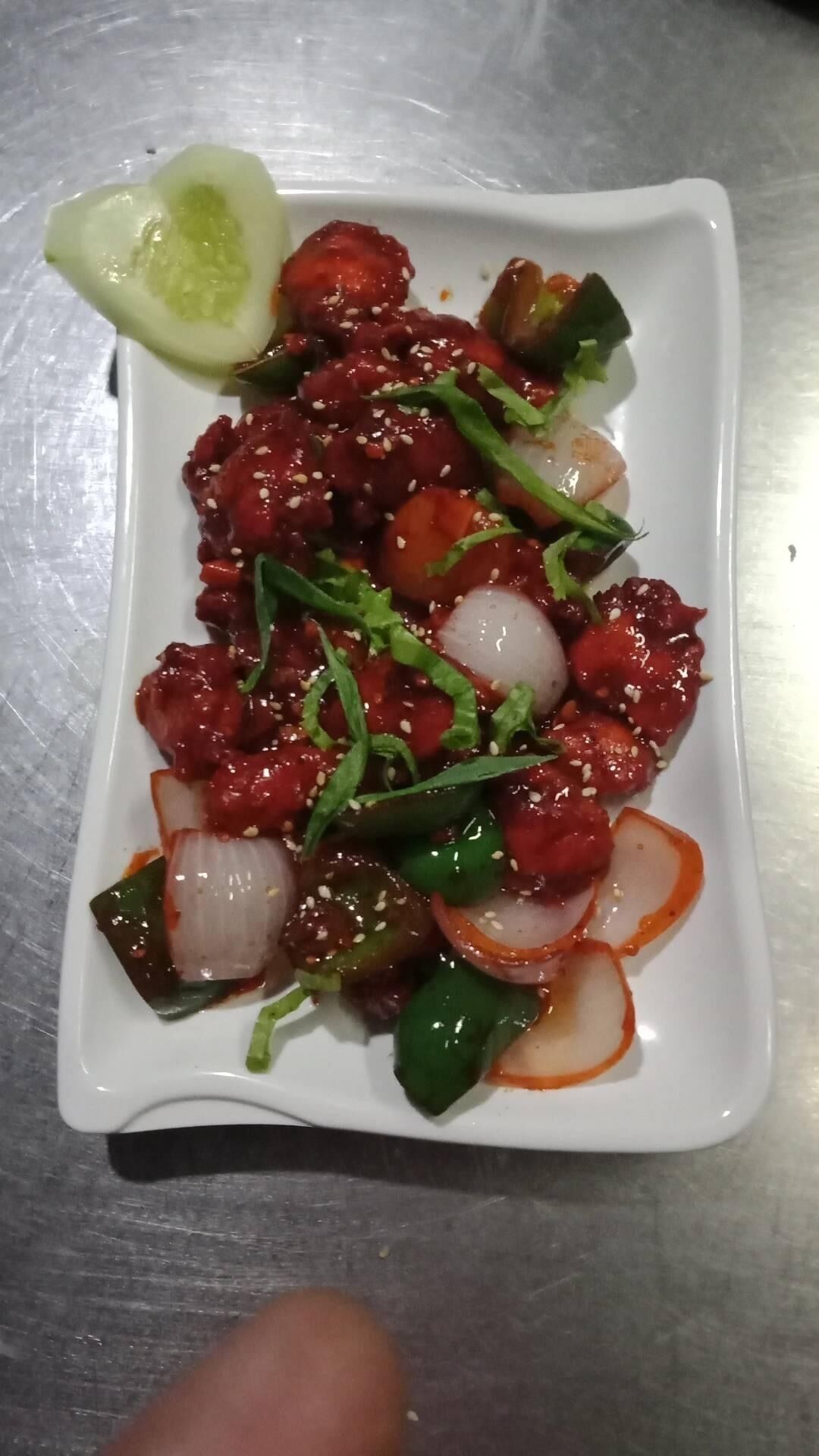 A white rectangular plate with a dish of stir-fried vegetables and meat, garnished with sesame seeds and green herbs, on a metallic surface.