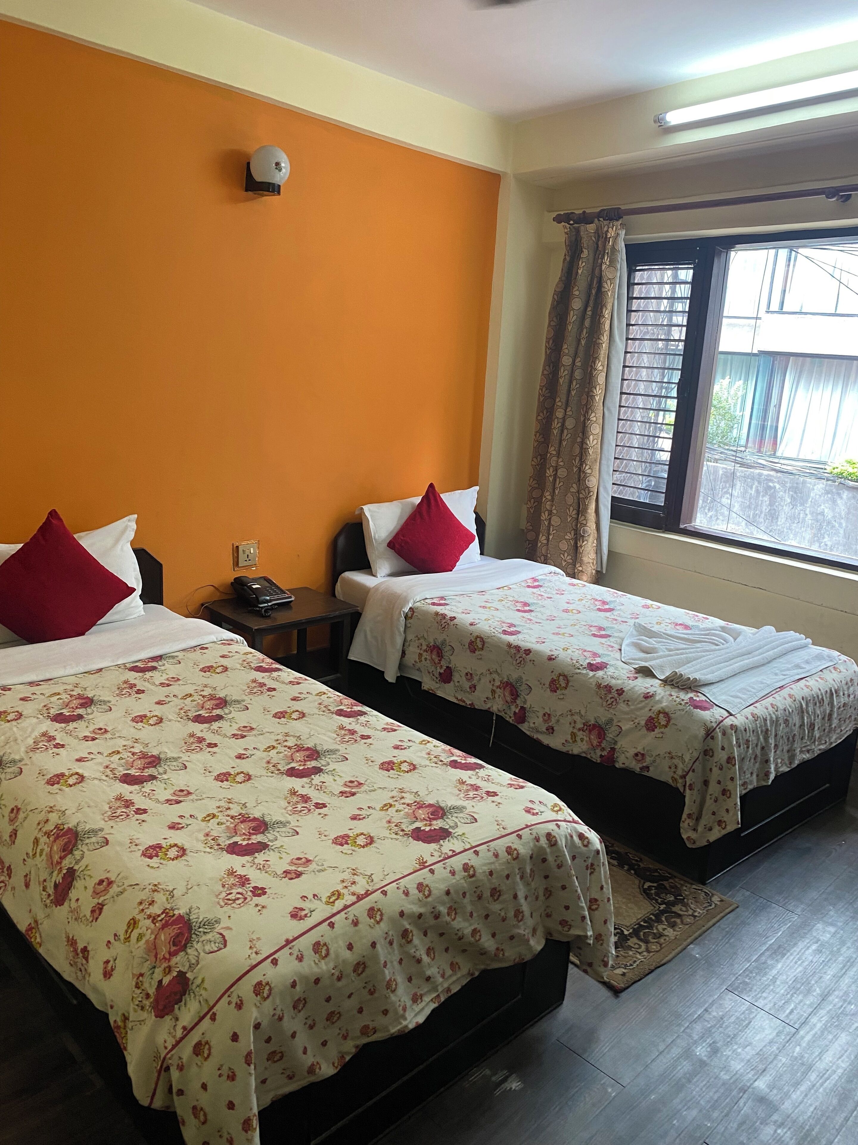 A hotel room with two beds, each with floral bedding and red pillows. There is a nightstand with a phone and a wall-mounted light. A window with curtains is visible, and a rug is on the floor.