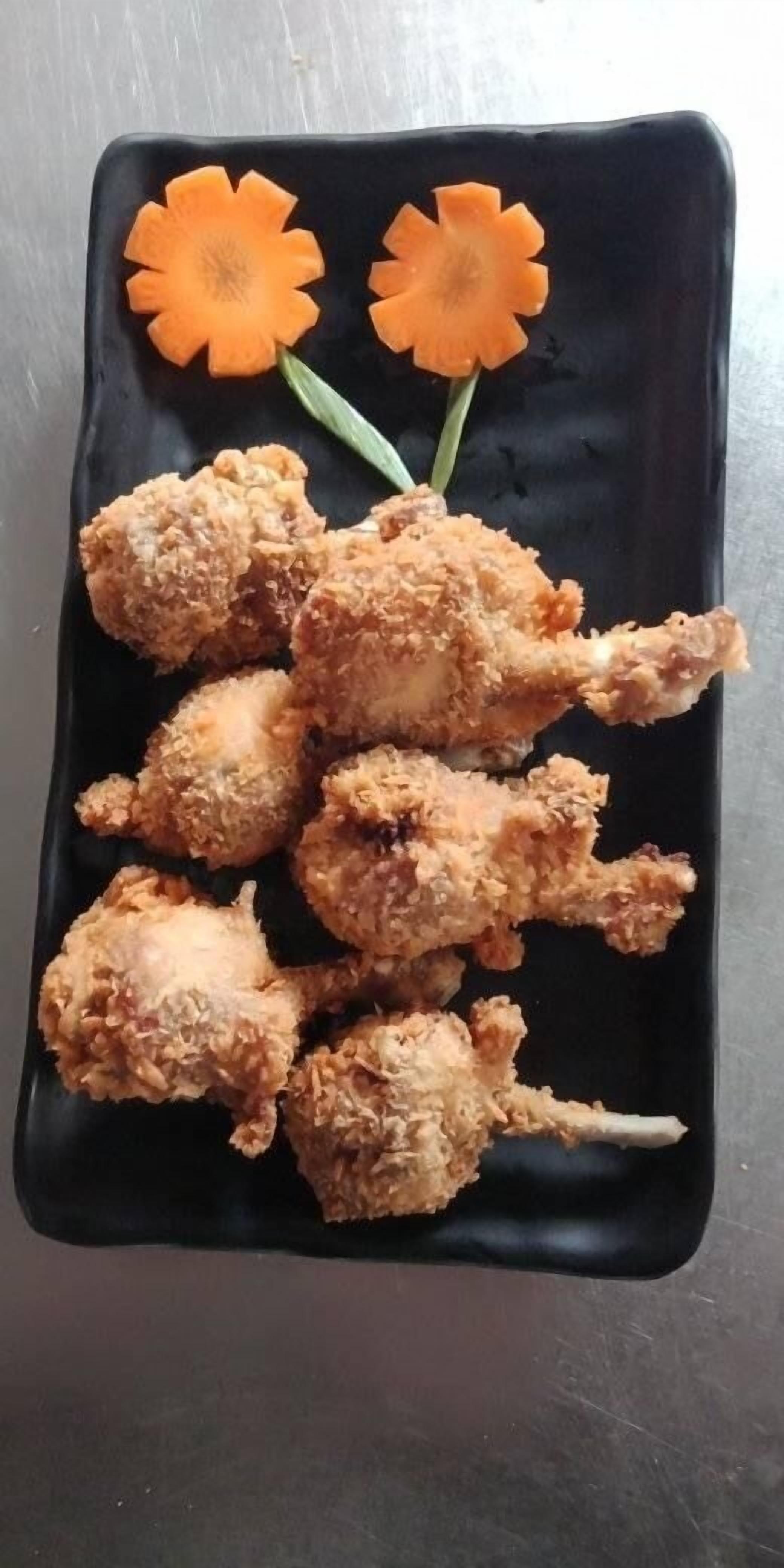 Une assiette rectangulaire noire sur laquelle on a disposé des morceaux de poulet frit et deux carottes sculptées.