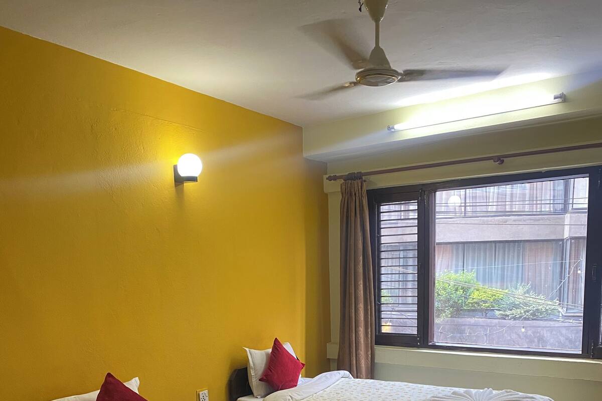 A hotel room with two beds, yellow walls, a ceiling fan, and a window with a view of greenery.