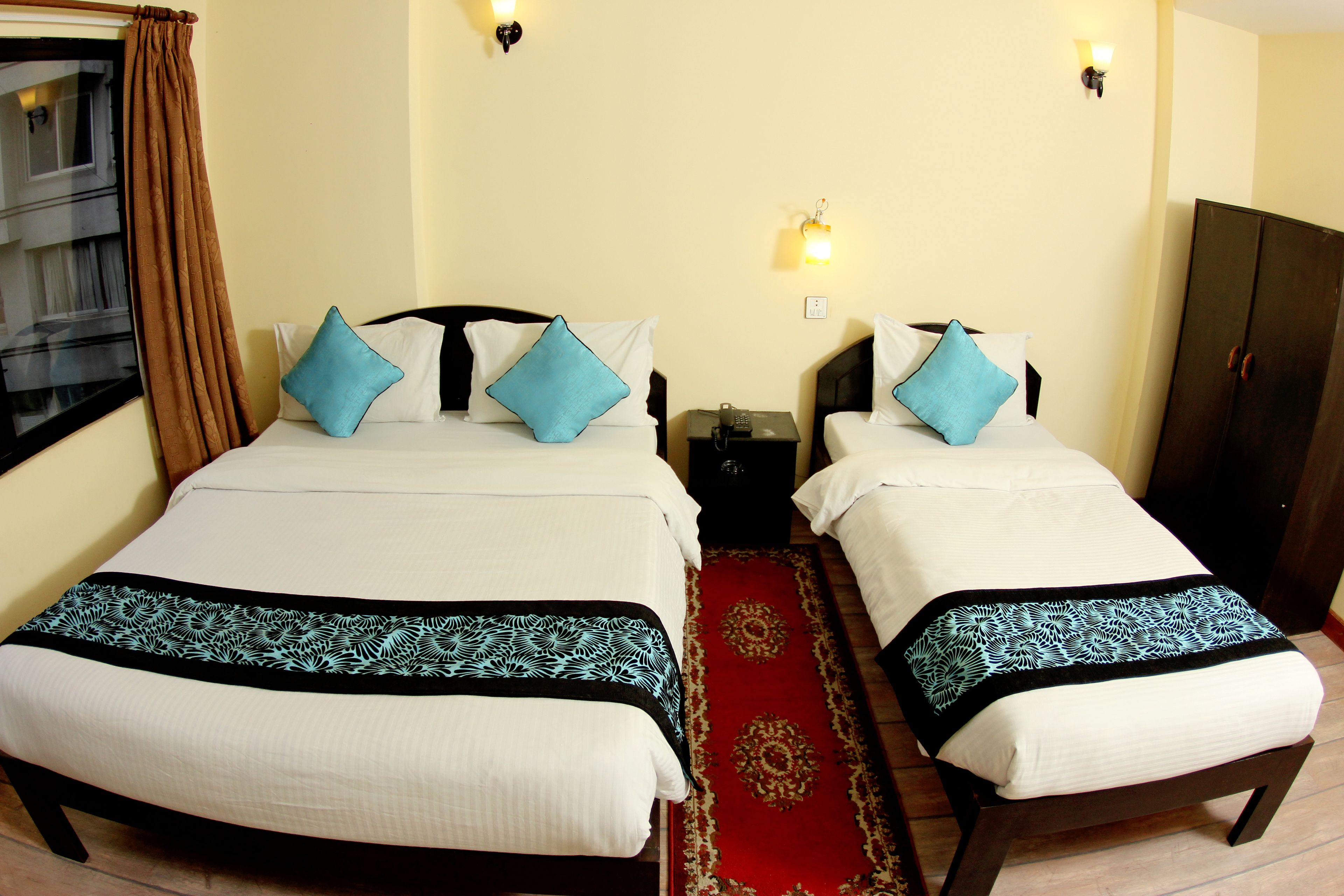 A double room with two beds, each with a white bedspread and a blue pillow. There is a red carpet runner between the beds, and a dark wooden wardrobe is visible to the right.