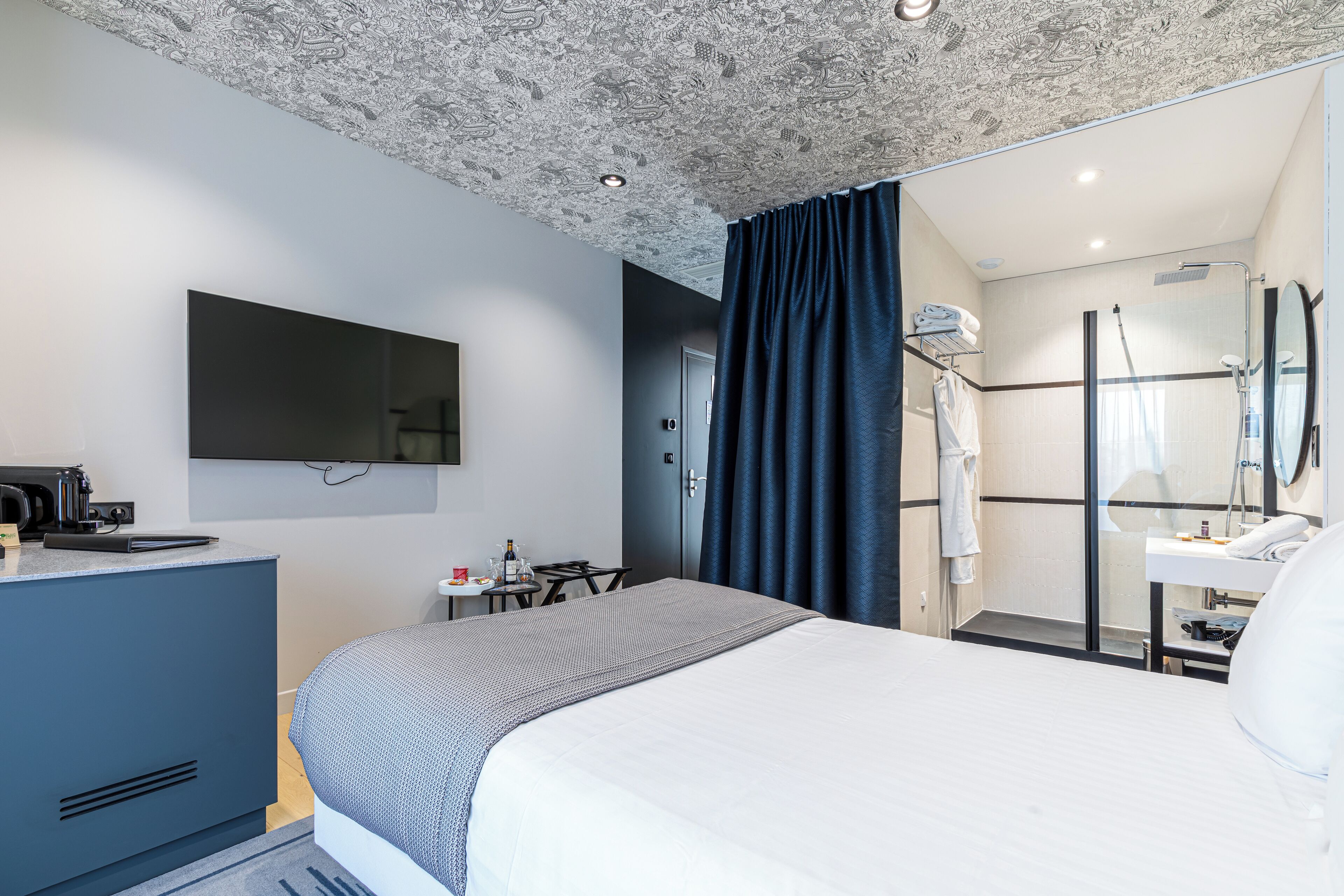 A modern hotel room with a large bed, a flat-screen TV, a desk with a kettle, and a shower area.