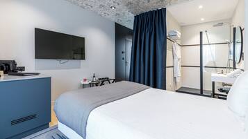 A modern hotel room with a large bed, a flat-screen TV, a desk with a kettle, and a shower area.