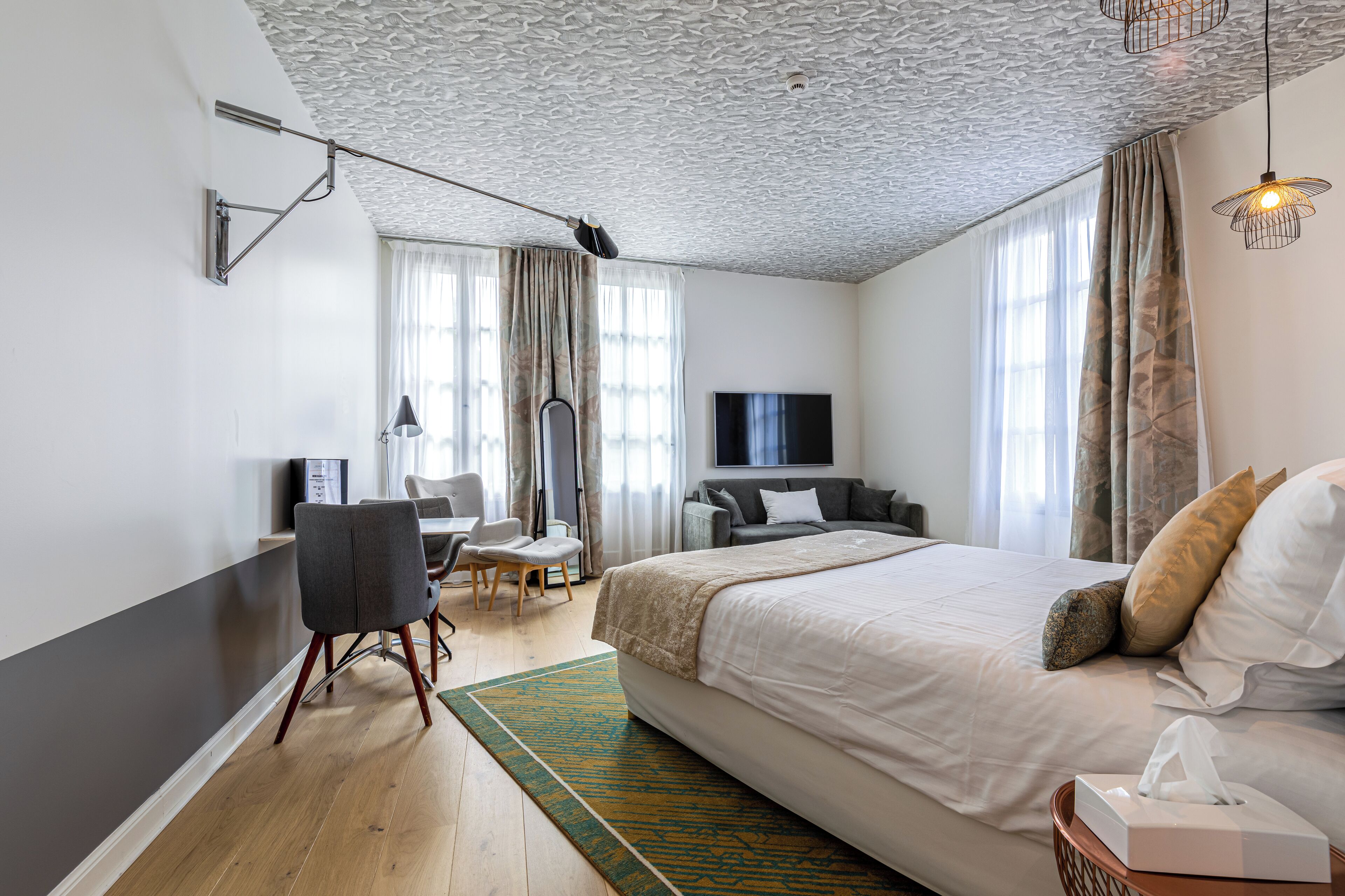 A modern hotel room with a large bed, a desk, a chair, a television, and a ceiling with a textured pattern.