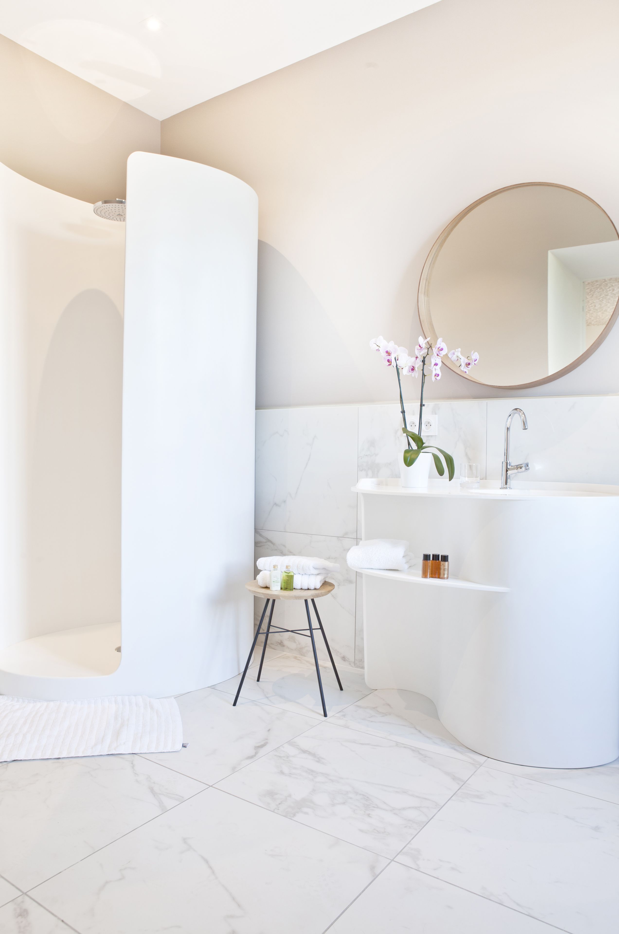 A modern bathroom with a white bathtub, a round mirror, a white sink, and a small stool with toiletries.