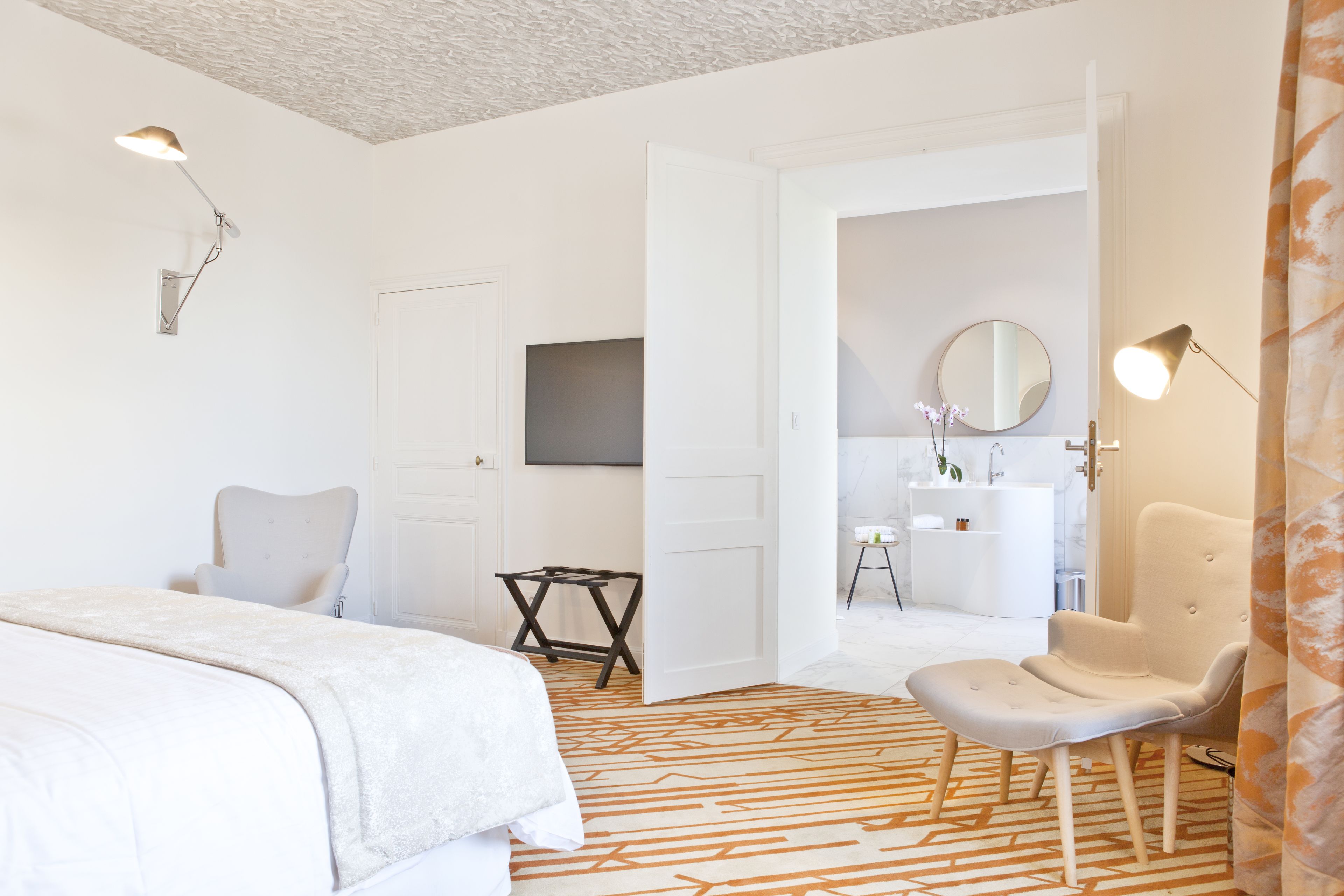 Ett modernt hotellrum med en säng, en stol, ett skrivbord, en tv och ett badrum som syns genom en öppen dörr.