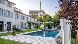 Une piscine entourée de chaises longues et de parasols, avec un jardin et un bâtiment historique en arrière-plan.