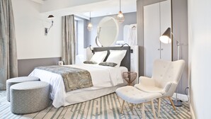 Une chambre d’hôtel moderne avec un grand lit, deux poufs, un fauteuil, un miroir rond et une armoire.