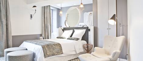 Una habitación de hotel moderna con una cama grande, dos otomanas, una silla, un espejo redondo y un armario.