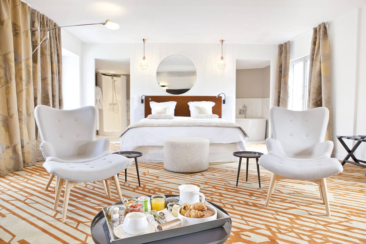 A hotel room with a large bed, two white armchairs, a breakfast tray, and a round mirror on the wall.