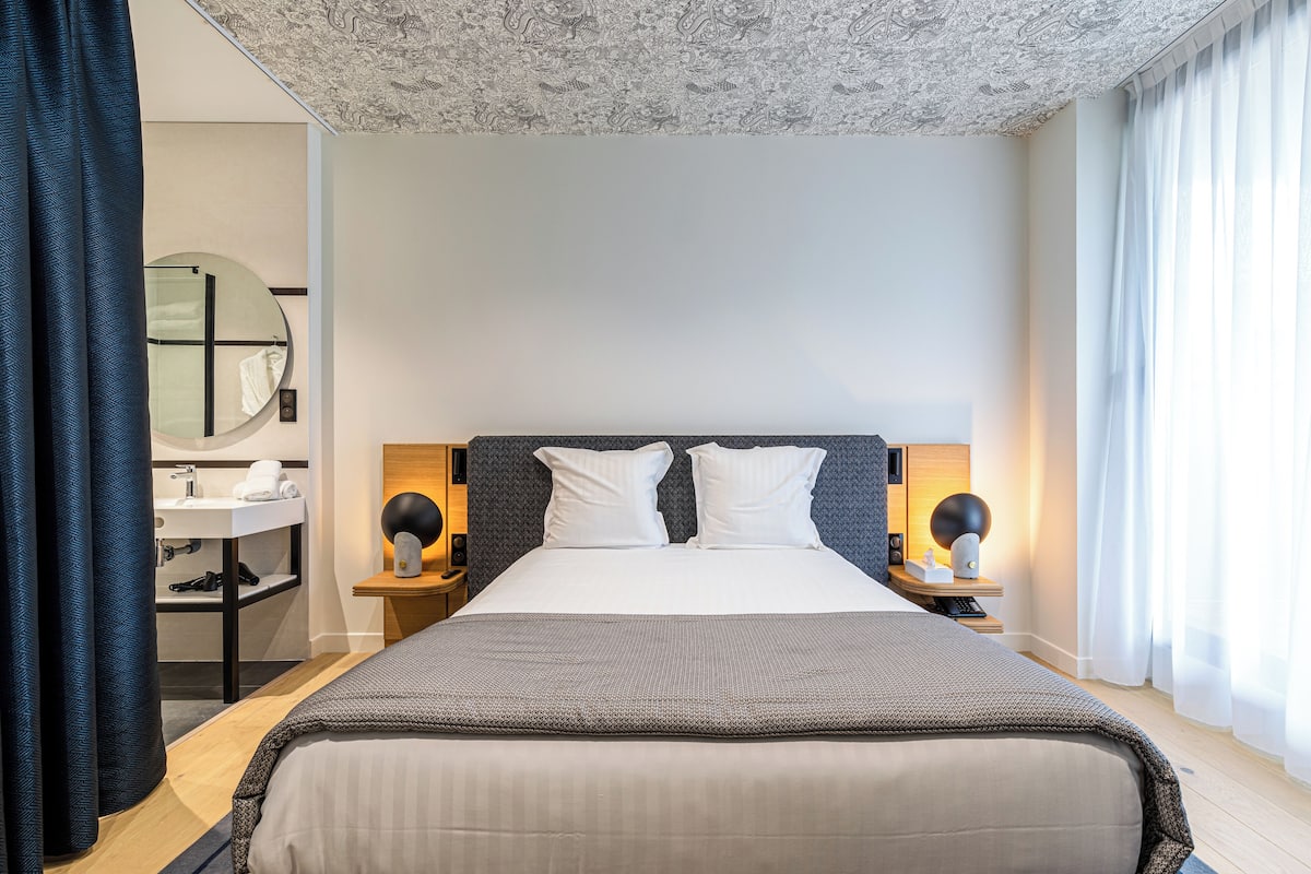A hotel room with a large bed, two bedside tables, a round mirror, a bathroom with a sink, and a patterned ceiling.