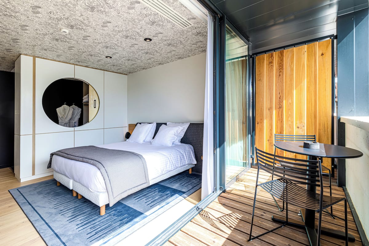 A modern bedroom with a bed, a round mirror, a small table, and a view of a wooden deck.