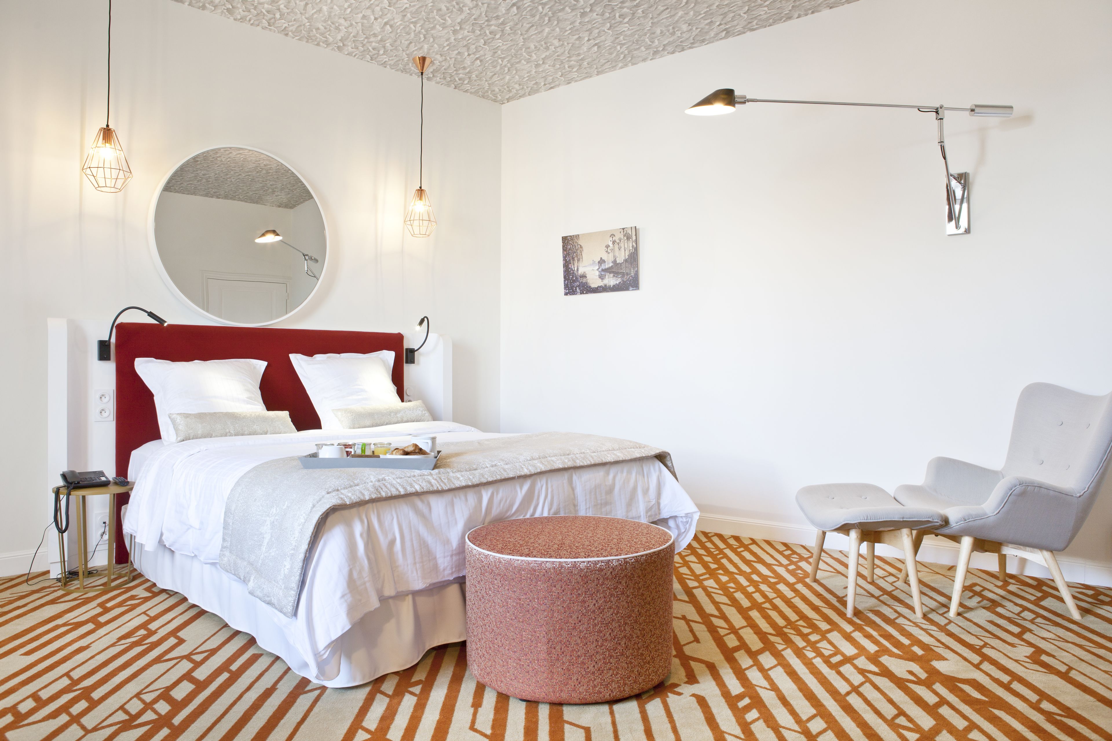 A modern bedroom with a red headboard, a round mirror, a bed with white bedding, a patterned rug, a round ottoman, a white chair, and a wall-mounted lamp.