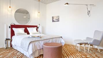 A modern bedroom with a red headboard, a round mirror, a bed with white bedding, a patterned rug, a round ottoman, a white chair, and a wall-mounted lamp.