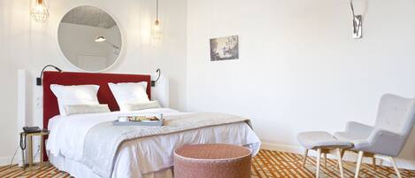 A modern bedroom with a red headboard, a round mirror, a bed with white bedding, a patterned rug, a round ottoman, a white chair, and a wall-mounted lamp.