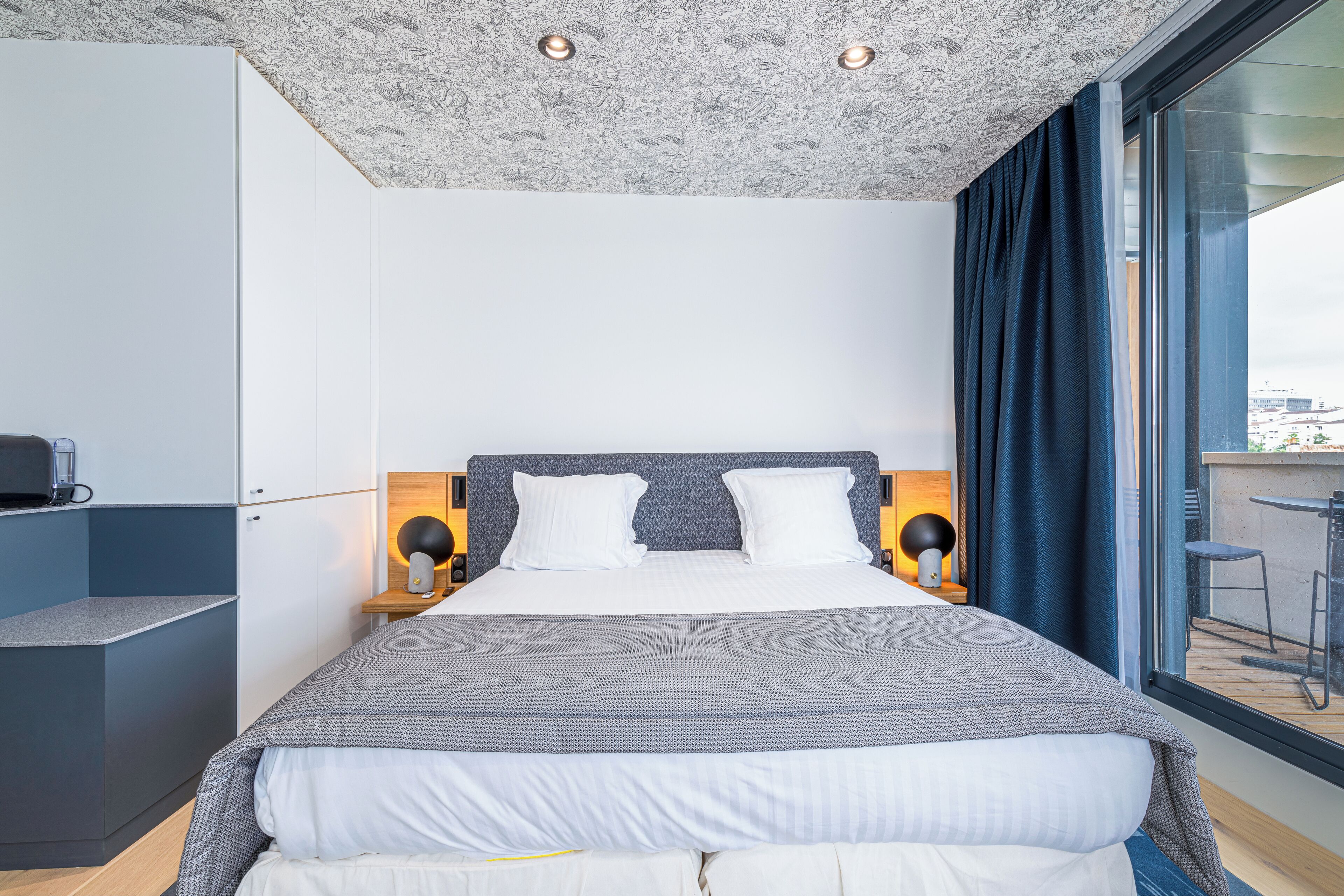 Habitación de hotel moderna con una cama grande, lámparas de noche, una mesita y vistas de la ciudad a través de un ventanal amplio.