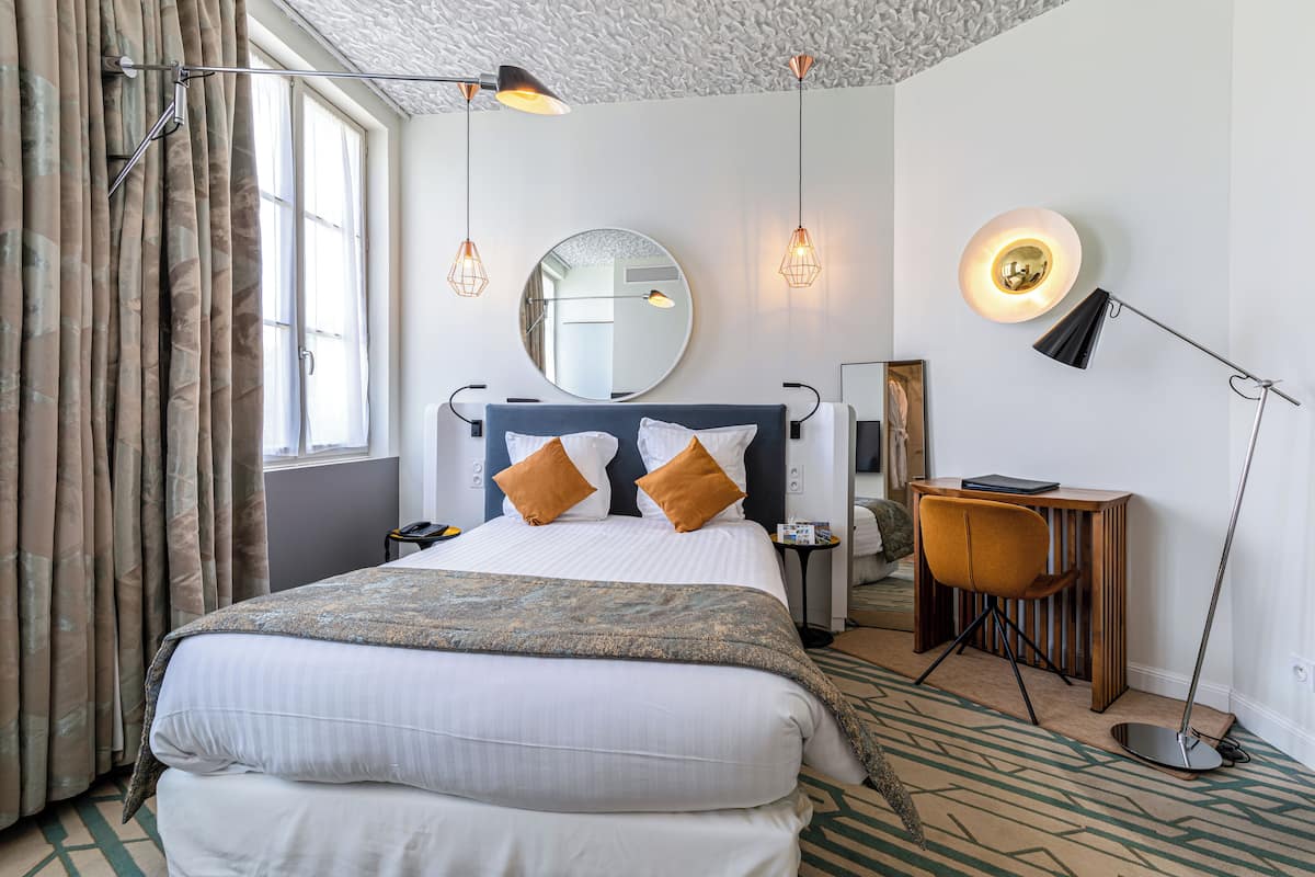 A hotel room with a large bed, a desk, a chair, a mirror, and a lamp.