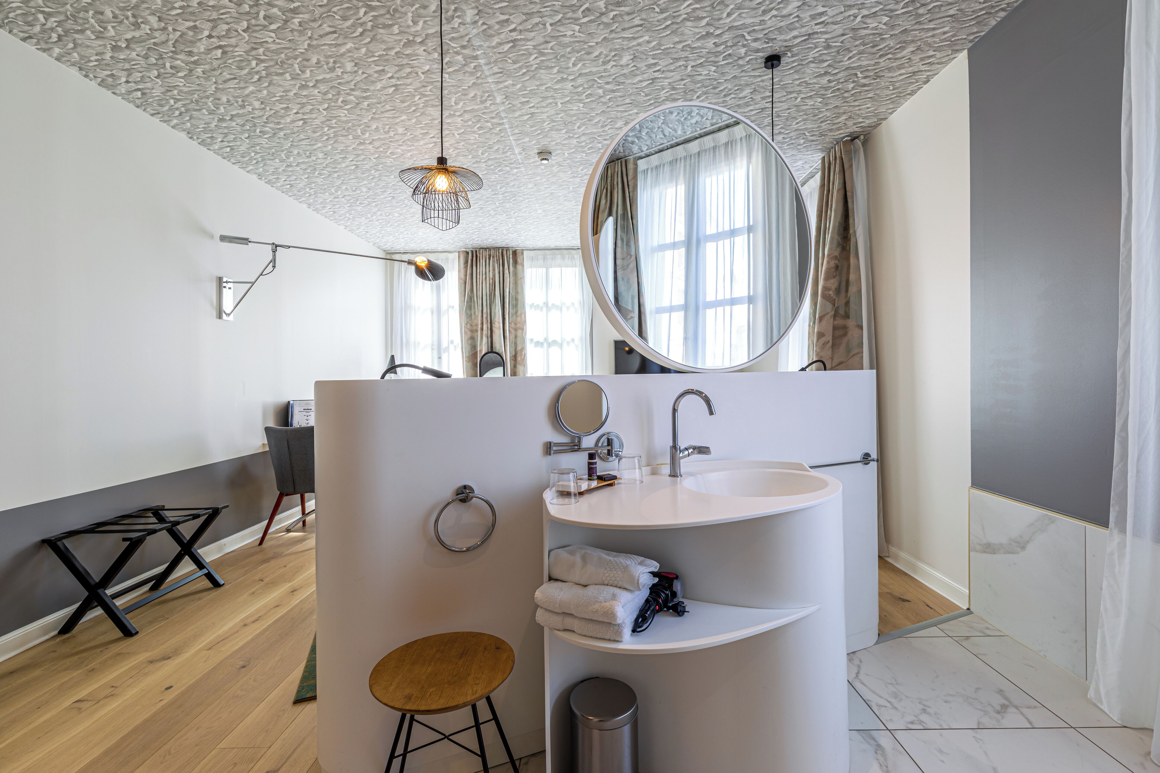 A modern bathroom with a white sink, a round mirror, and a wooden stool.