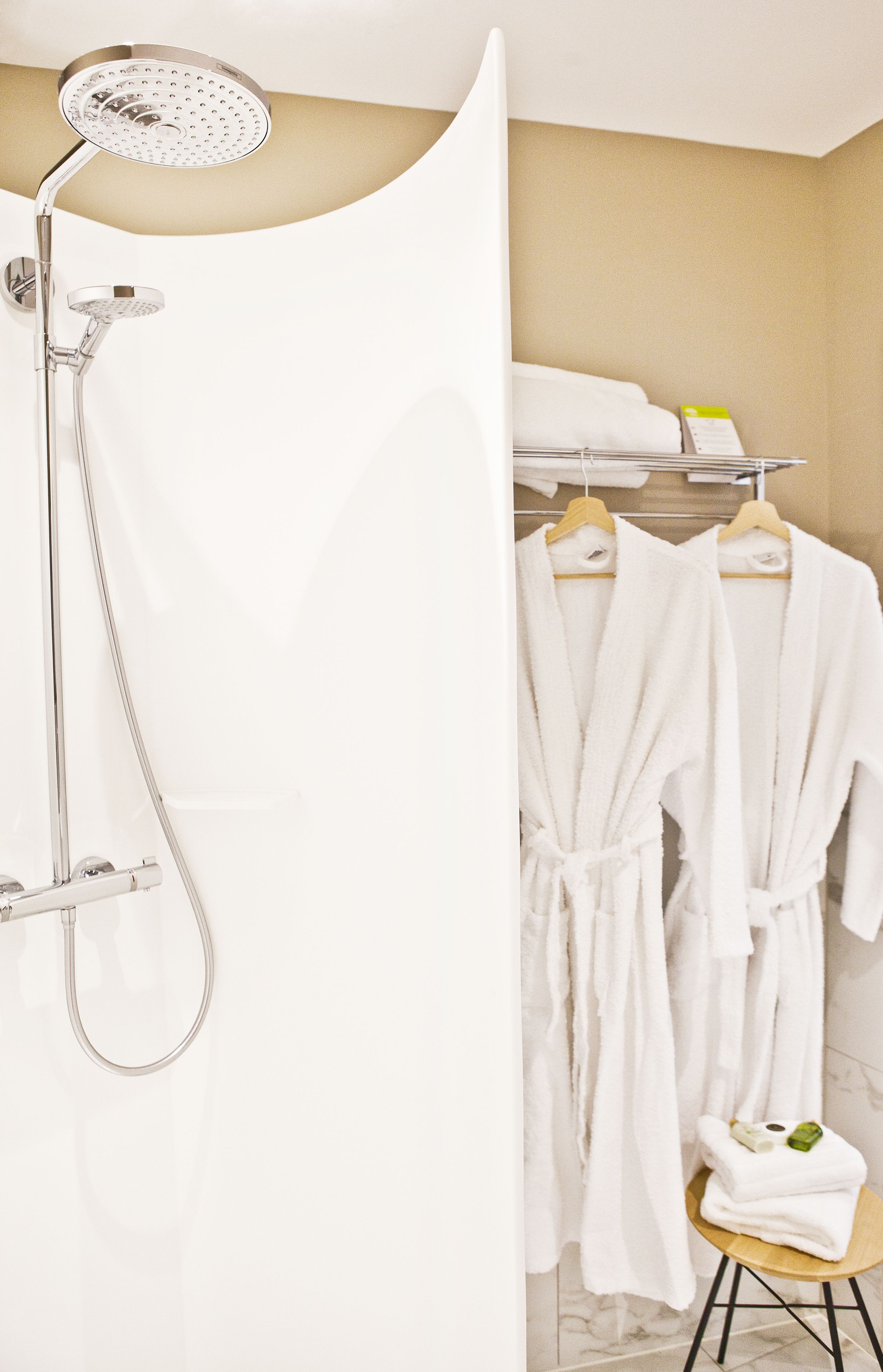 A bathroom with a shower, two white robes, a towel rack, and a small stool with towels.
