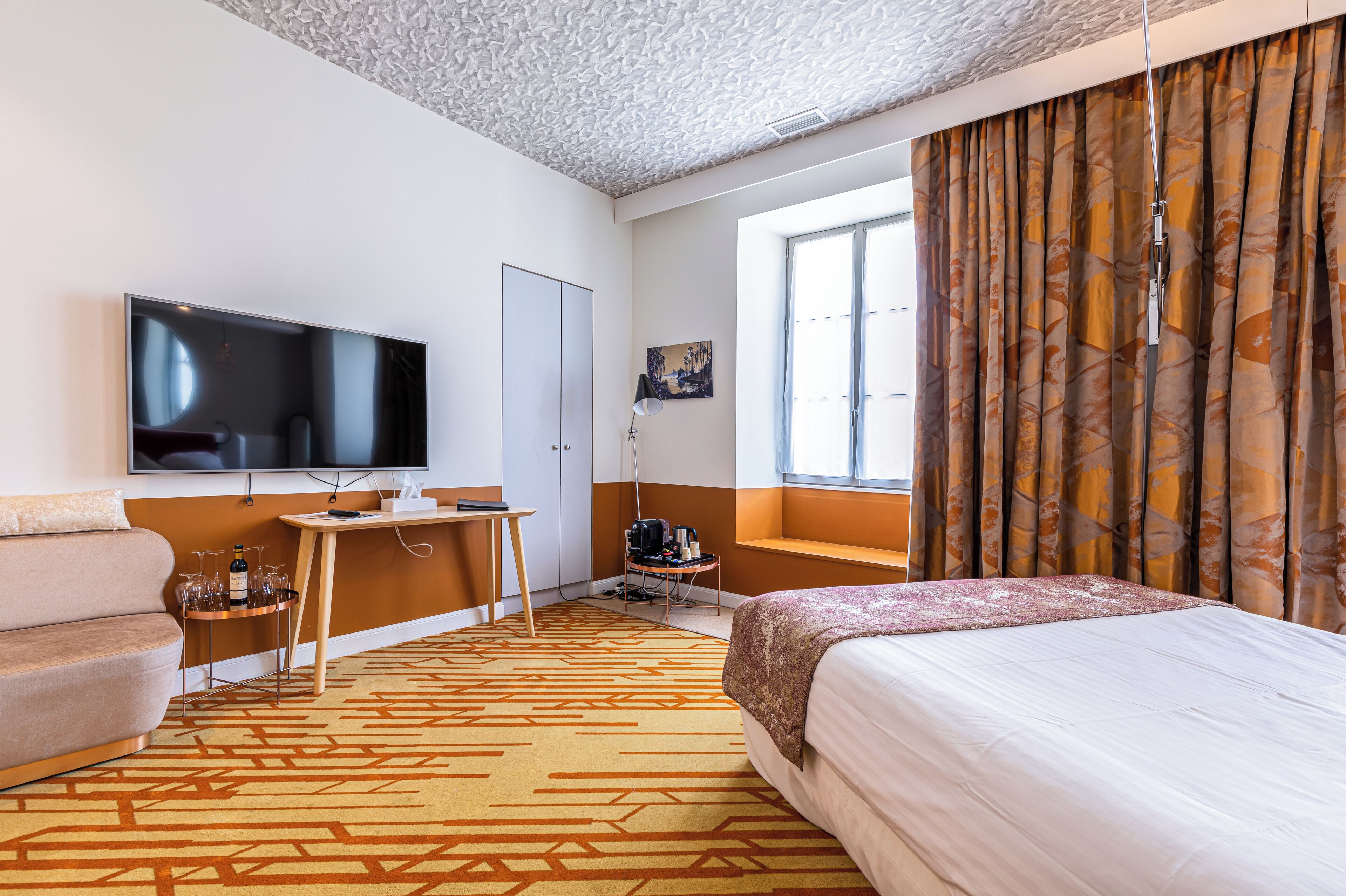 A hotel room with a bed, a desk with a computer, a TV, and a window with curtains.