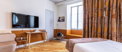 A hotel room with a bed, a desk with a computer, a TV, and a window with curtains.