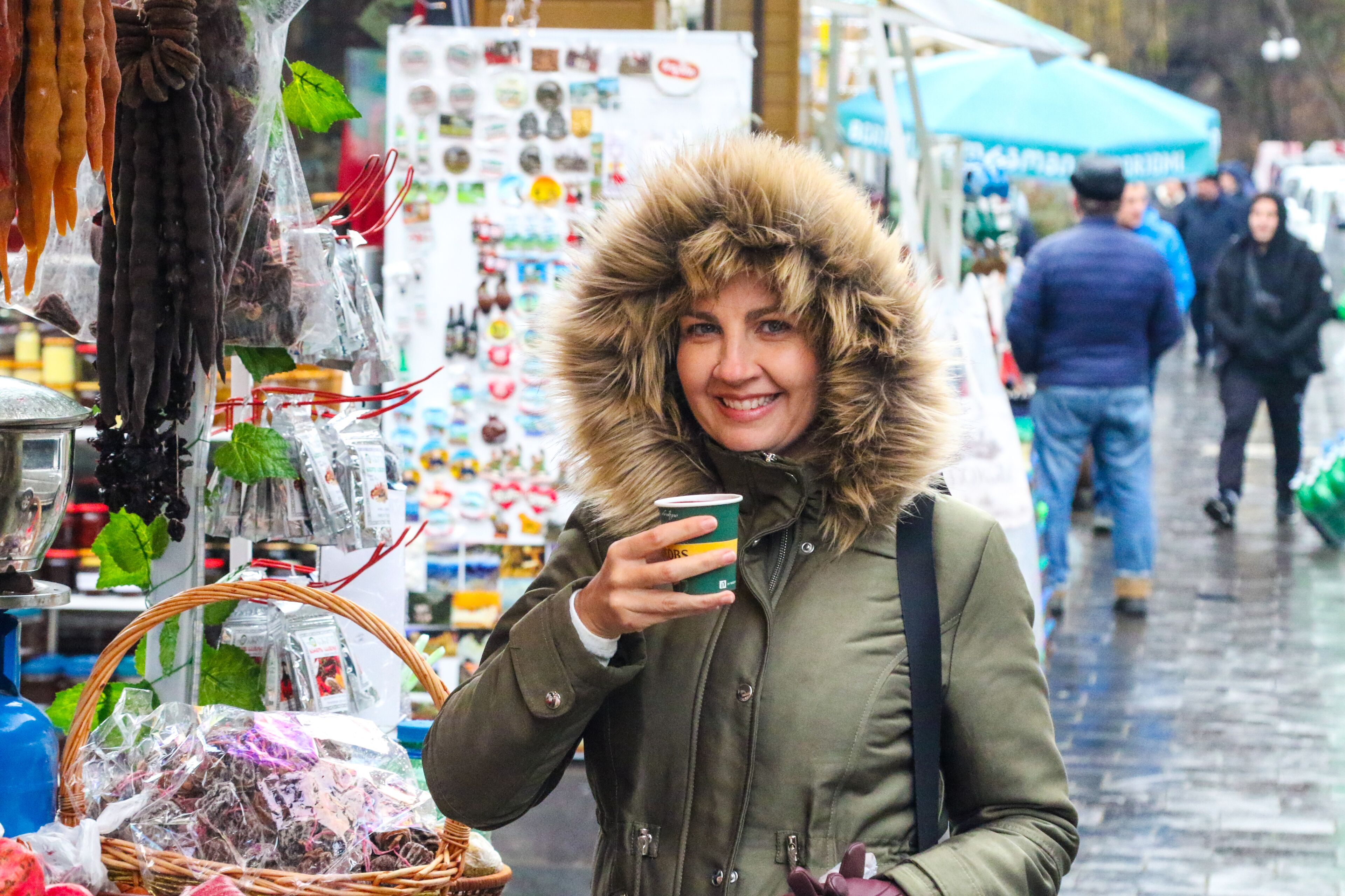 A person in a fur-lined coat holding a cup, standing in a market with various items for sale.