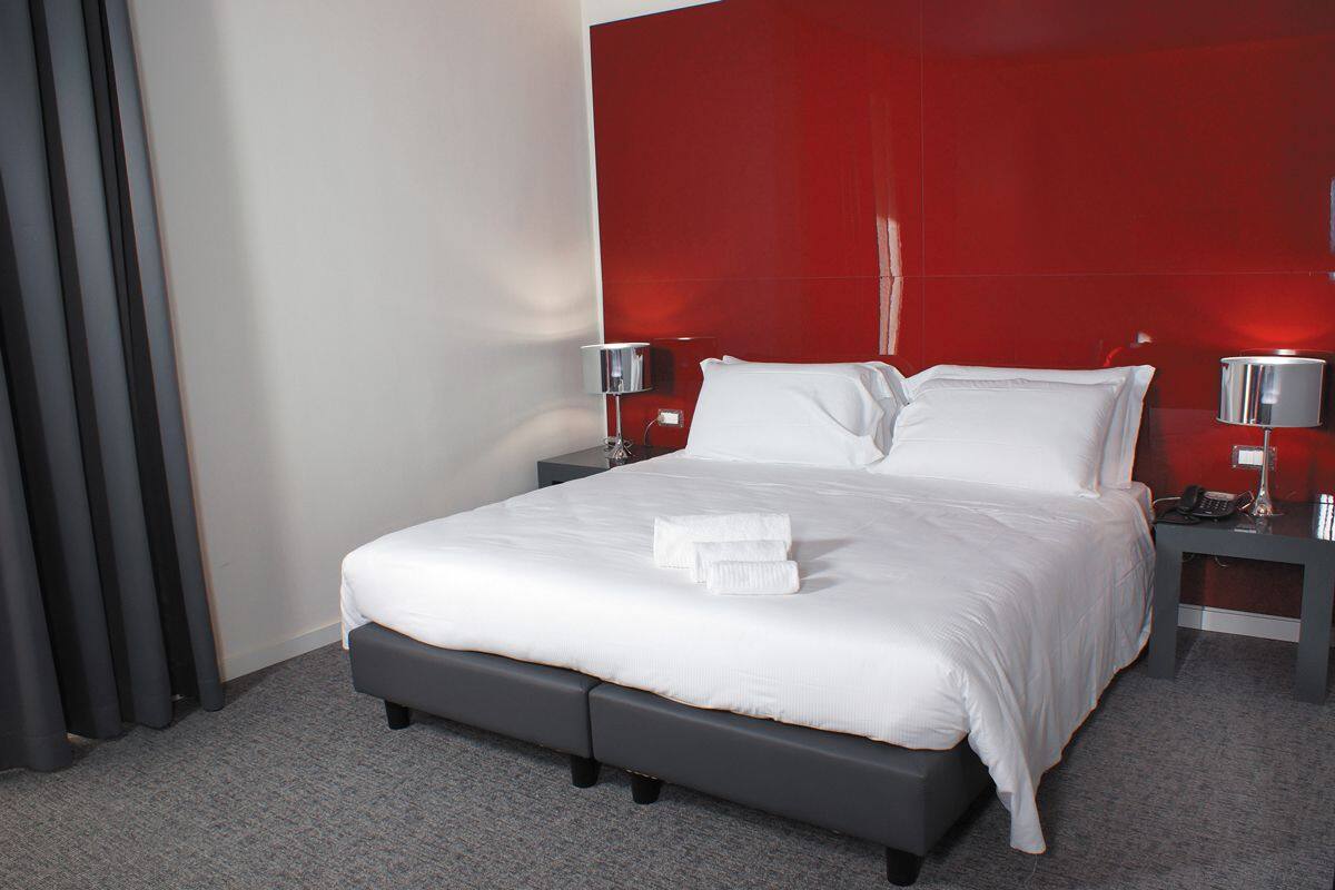 A modern hotel room with a large bed, grey padded headboard, white linens, two bedside tables with lamps, and a red accent wall.