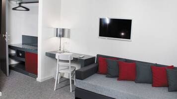 Quarto moderno com sofá cinza, almofadas vermelhas e cinzas, uma escrivaninha pequena com cadeira, televisão na parede e um guarda-roupa com cabideiro.