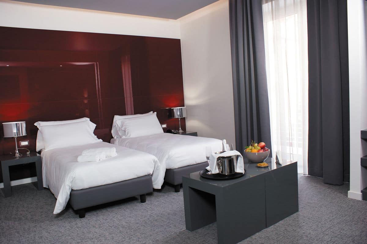A hotel room with two beds, a desk with a bowl of fruit, and a large window with curtains.