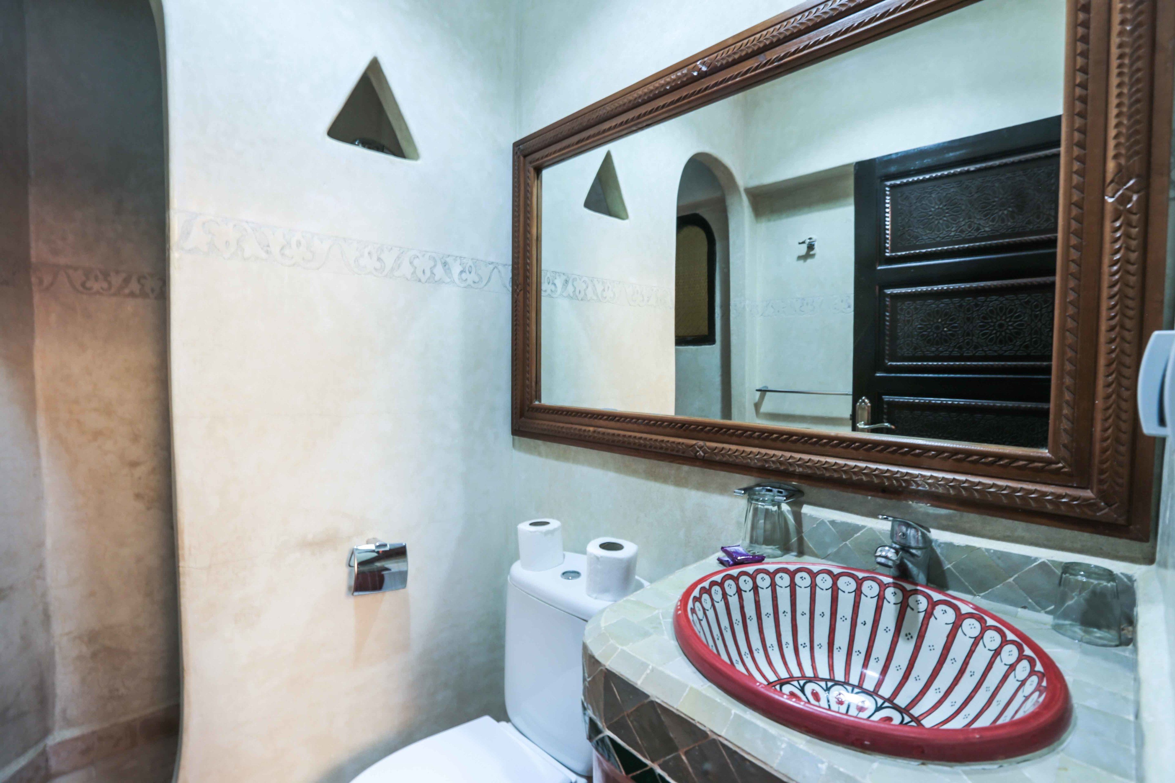 Un baño con inodoro blanco, lavamanos de cuenca roja y espejo con marco de madera.